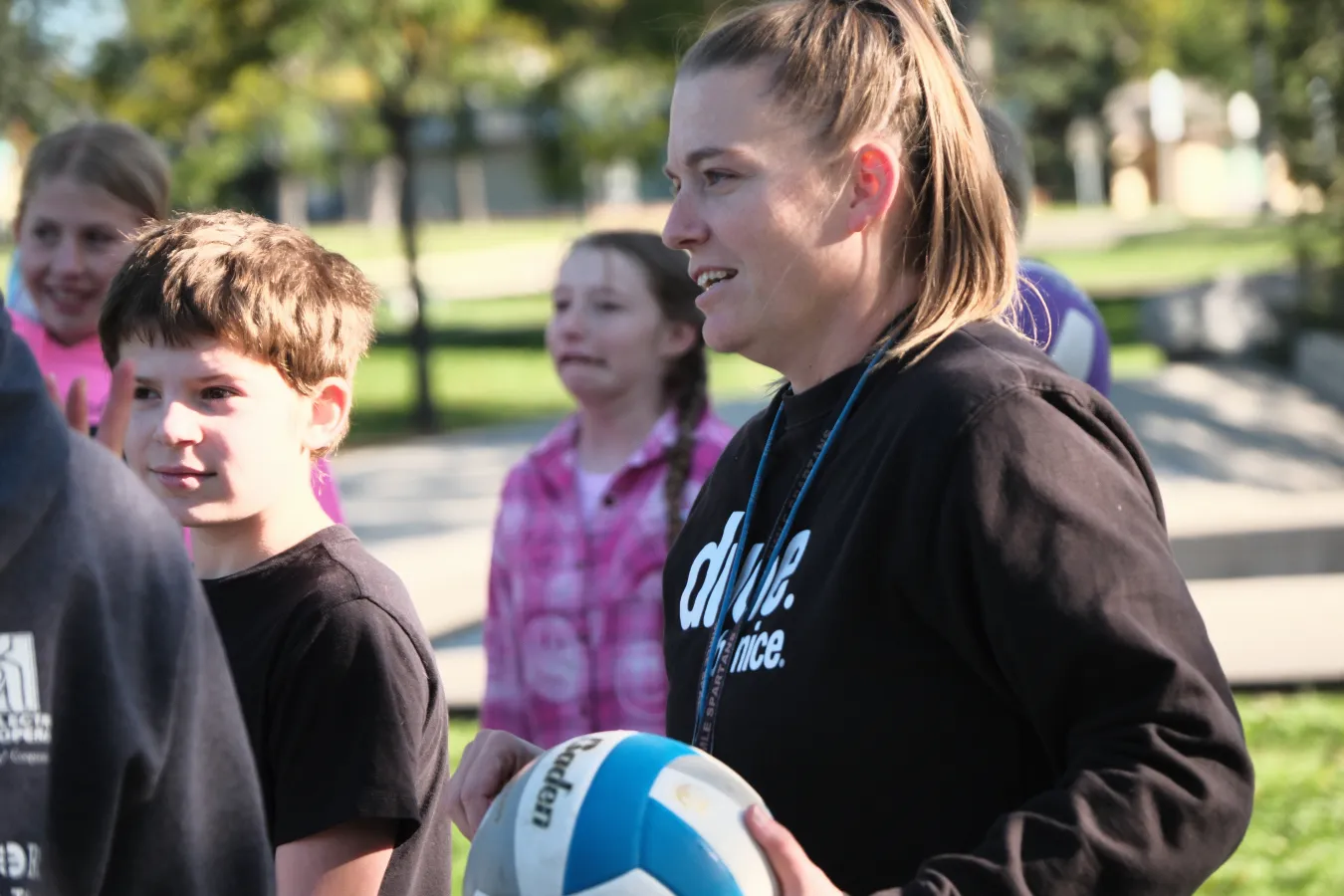 A teacher holds a volleyball while standing with students outdoors, giving instructions during a physical education activity on a sunny day.