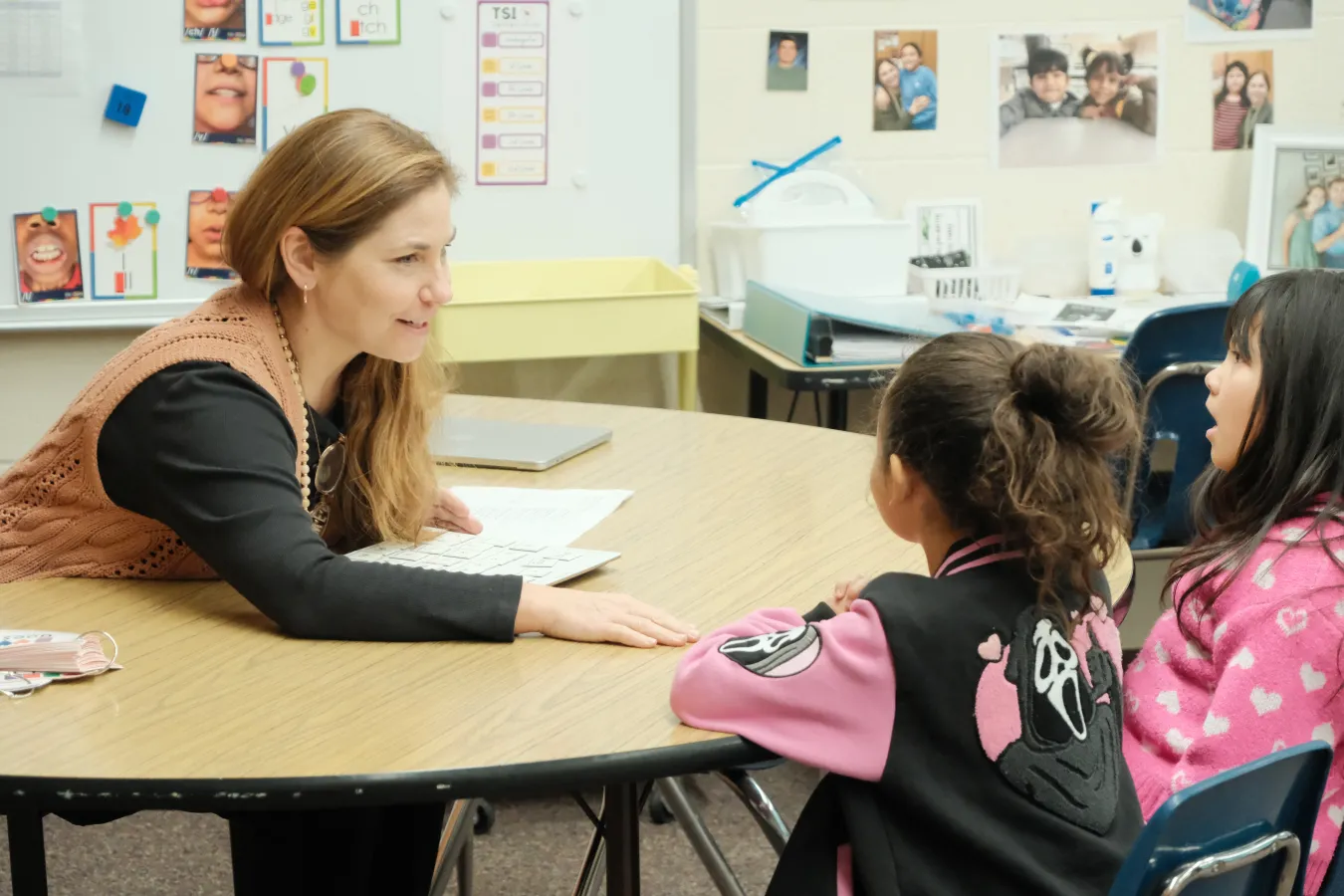 A teacher sits at a round table speaking with two students during a small-group reading lesson in an elementary classroom.