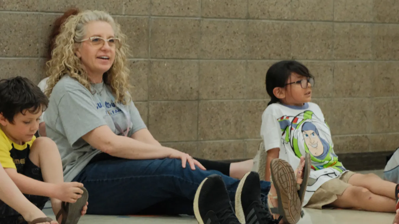 2027 North Dakota United Education Support Professional of the Year Dawn Ressler sits on a gym floor with elementary students during a physical therapy activity at Mandan Public Schools. Ressler smiles as she engages with the children during a playful movement exercise.
