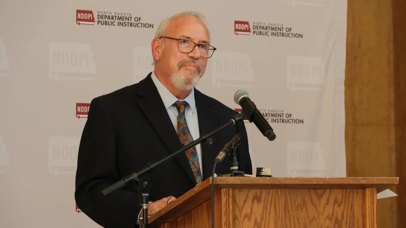 North Dakota United President Nick Archuleta stands at a podium with microphones in front of a North Dakota Department of Public Instruction backdrop.