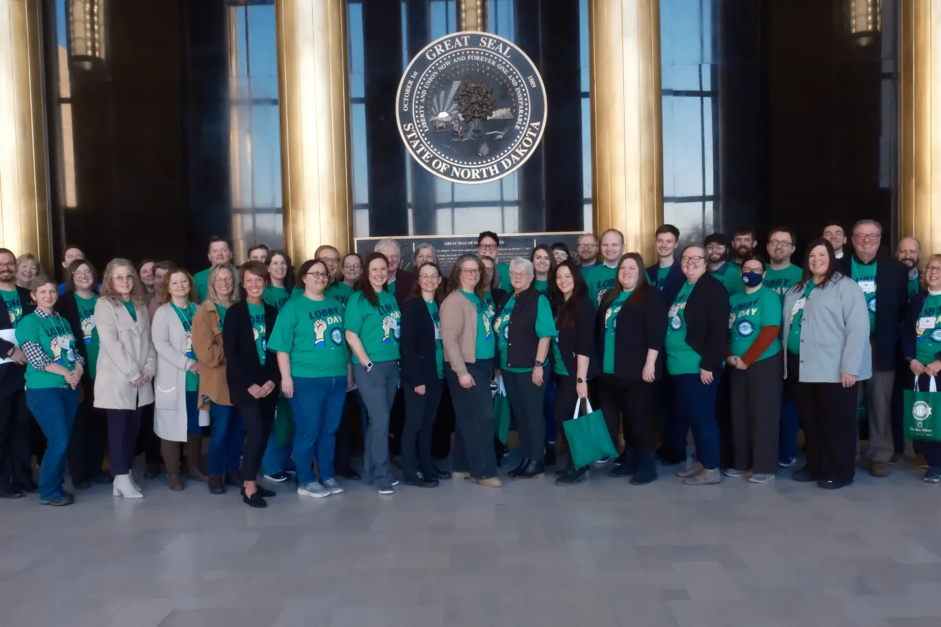 A large group of people — some wearing green T-shirts printed with "Lobby Day" and the ND United logo — stand together inside Memorial Hall of the state Capitol building.