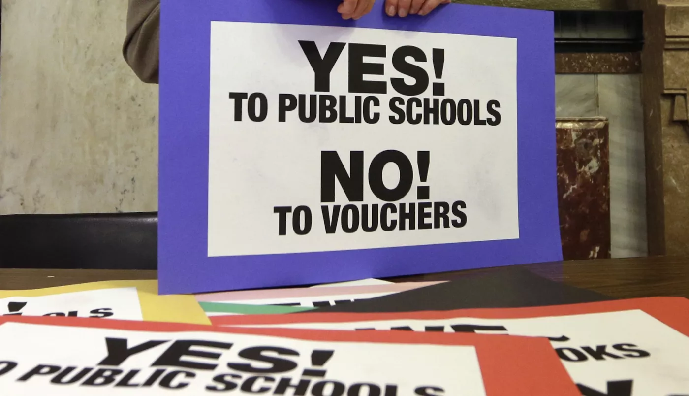 A sign with a bright purple border is held on a table just in frame. In black text against a white background it says: "YES! to Public Schools. NO! to Vouchers." The table is filled with more signs that say the same thing with different colored borders.