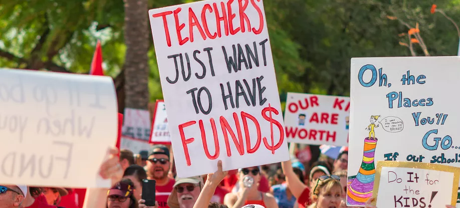 A sign is held up at a protest that says "Teachers just want to have funds" with the "s" in "funds" made to look like a dollar sign. Other signs near by say, "Do it for the kids!" and "Our kids are worth it!" Another sign has a drawing that shows the cover of the Doctor Seuss book, "Oh, the Places You'll Go."