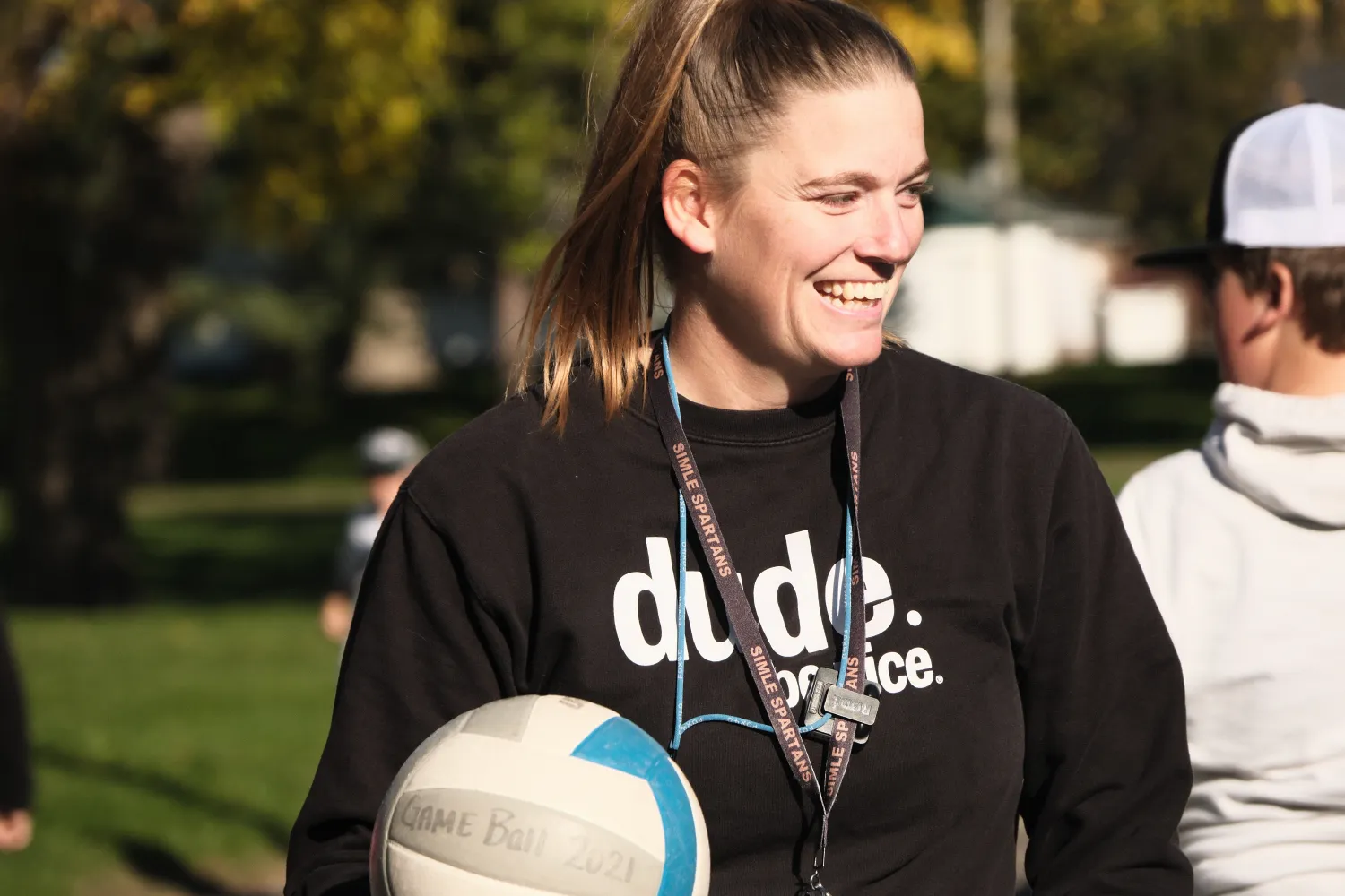 A teacher smiles while holding a volleyball during an outdoor PE class, with students and green space visible in the background.