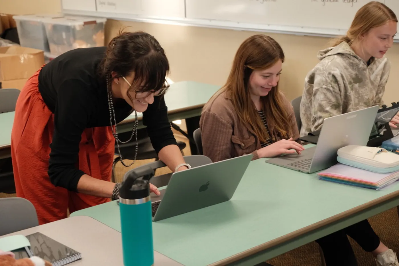 A teacher leans over a table to assist two students working on laptops, smiling as she offers guidance in a collaborative classroom setting.
