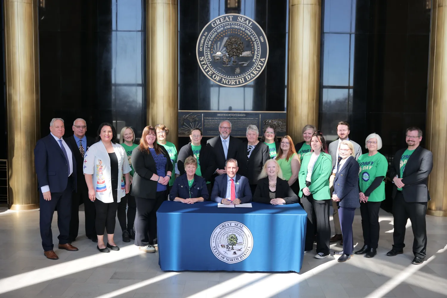A large group of people — some wearing green T-shirts printed with "Lobby Day" and the ND United logo — stand behind three individuals sitting at a table with a blue tablecloth and the official seal of the state of North Dakota, inside Memorial Hall of the state Capitol building.