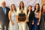 Six individuals standing in a row, posing for photo, with woman in middle holding North Dakota Teacher of the Year trophy.