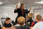 A teacher stands beside a group of students seated at desks, raising both hands while holding a sheet of paper as she speaks. Students around her are writing and listening during a classroom lesson.