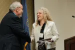 A woman holding a certificate smiles as she shakes hands with a man during an award presentation beside a podium and state flag.