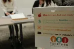 A box labeled with campaign messaging sits in the foreground as Secretary of State staff sort petition sheets in the background.