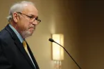 North Dakota United President Nick Archuleta speaks at a podium during the 2026 North Dakota United Delegate Assembly in Bismarck. He is shown in profile wearing a dark suit, light blue shirt and gold tie, speaking into a microphone.