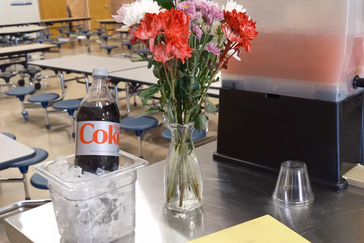 Flowers in a vase and a two-liter bottle of Diet Coke in a container of ice, sit on a table in a school cafeteria.