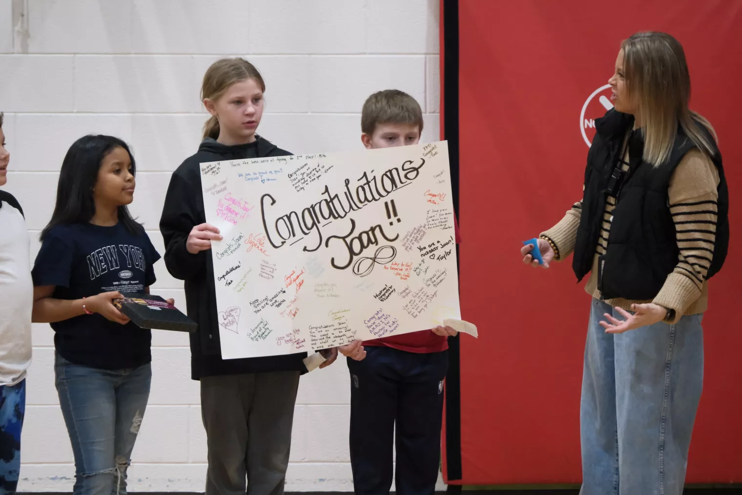 A group of people stand together indoors, holding a large poster with handwritten messages that reads "Congratulations Joan!.