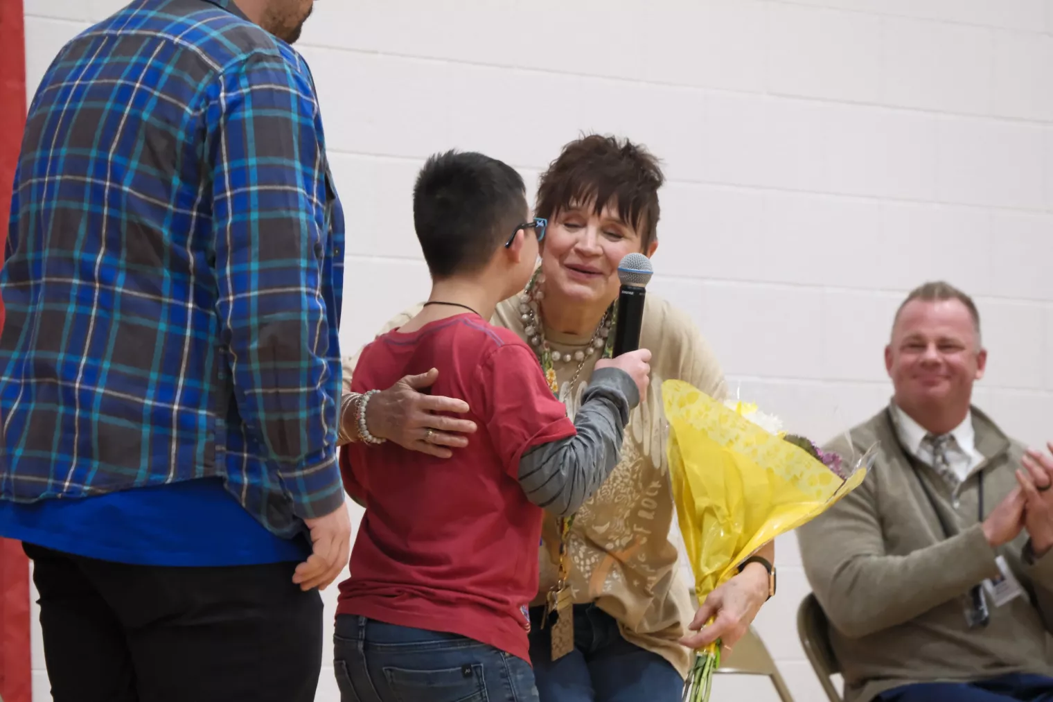A boy in a red shirt stands with a woman holding yellow flowers and a microphone. Another person in a plaid shirt stands nearby. A seated man claps in the background.