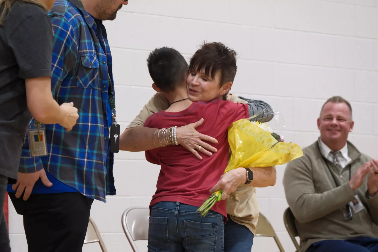 A student in a red shirt hugs a woman holding yellow flowers in a gymnasium. Other people stand and sit around them, clapping and smiling. The background features a white brick wall and foldable chairs.