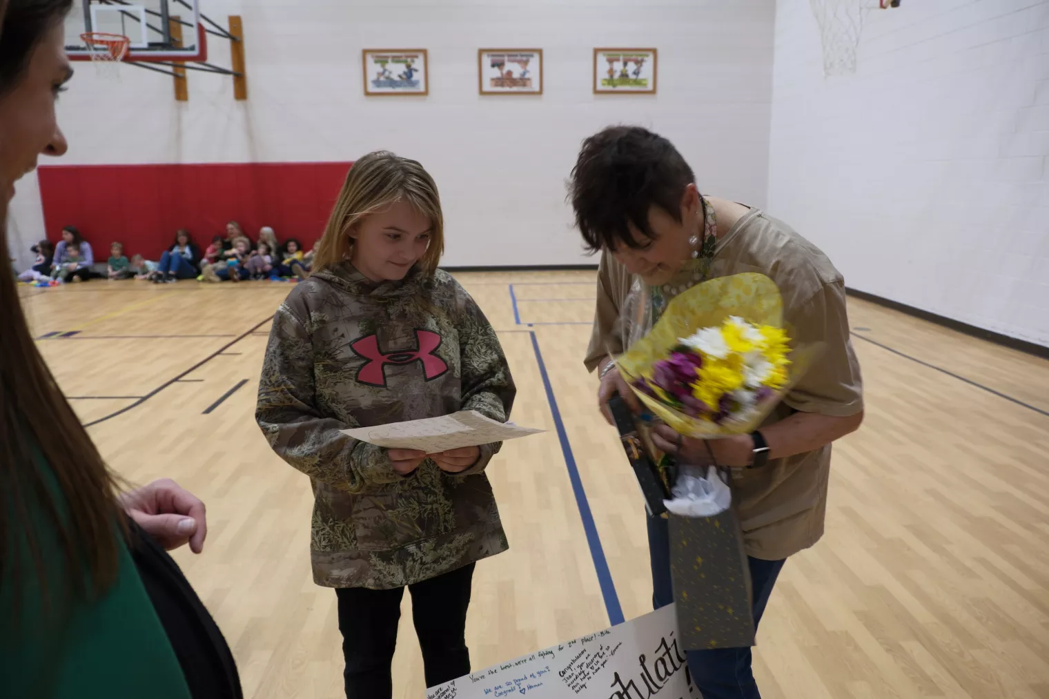 A young girl in a camouflage hoodie smiles while holding a paper in a gymnasium. Beside her, a woman with short hair and glasses is presenting a bouquet of yellow and purple flowers. A group of people are seated in the background.
