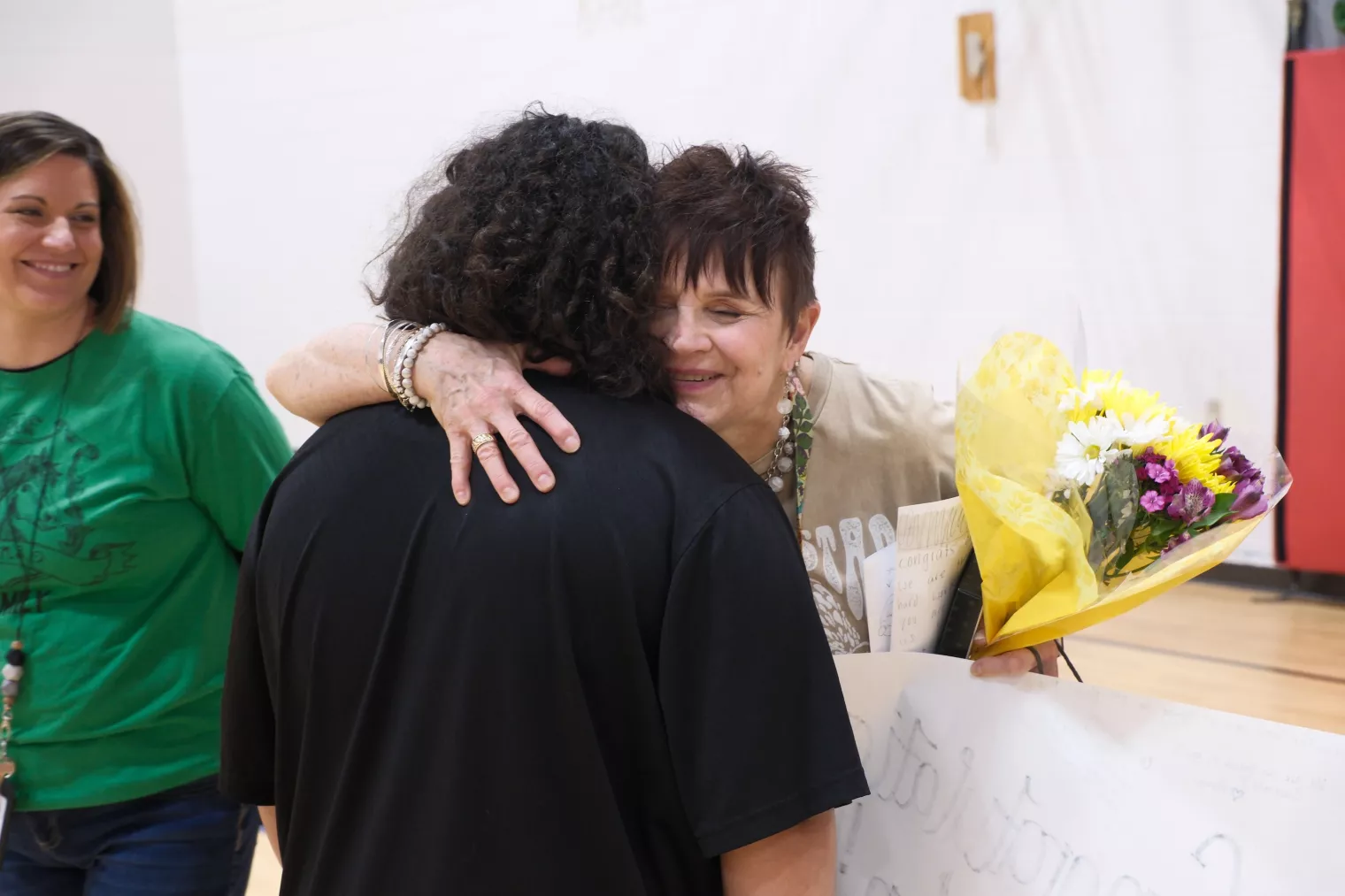 A person with short hair hugs a person with curly hair in a gymnasium. The short-haired person holds a bouquet of flowers and a large piece of paper. Another person in a green shirt stands nearby, smiling.
