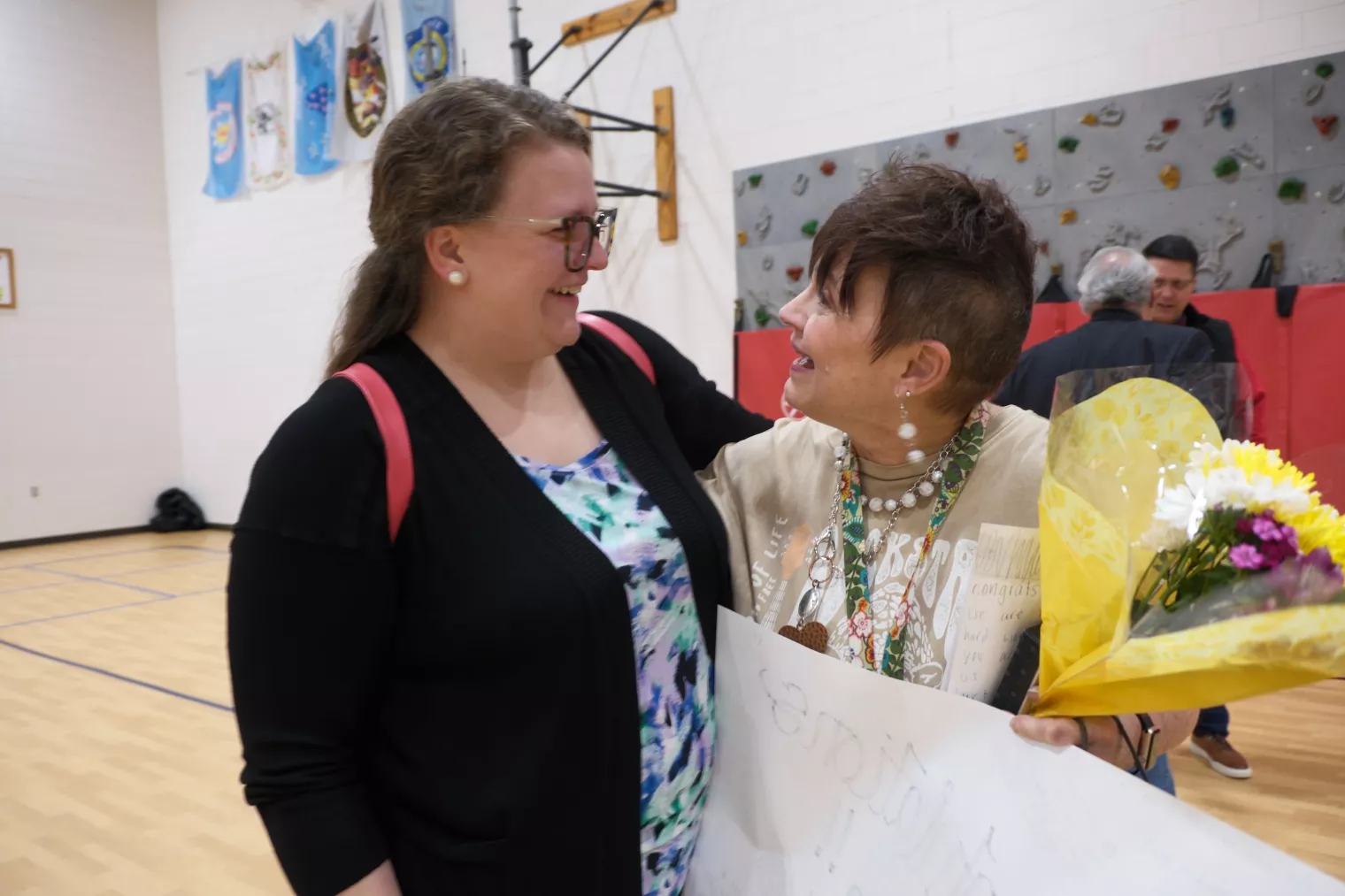 Two women stand in a gymnasium, smiling and embracing. One holds a bouquet of flowers and a large card. Colorful banners hang on the wall behind them.
