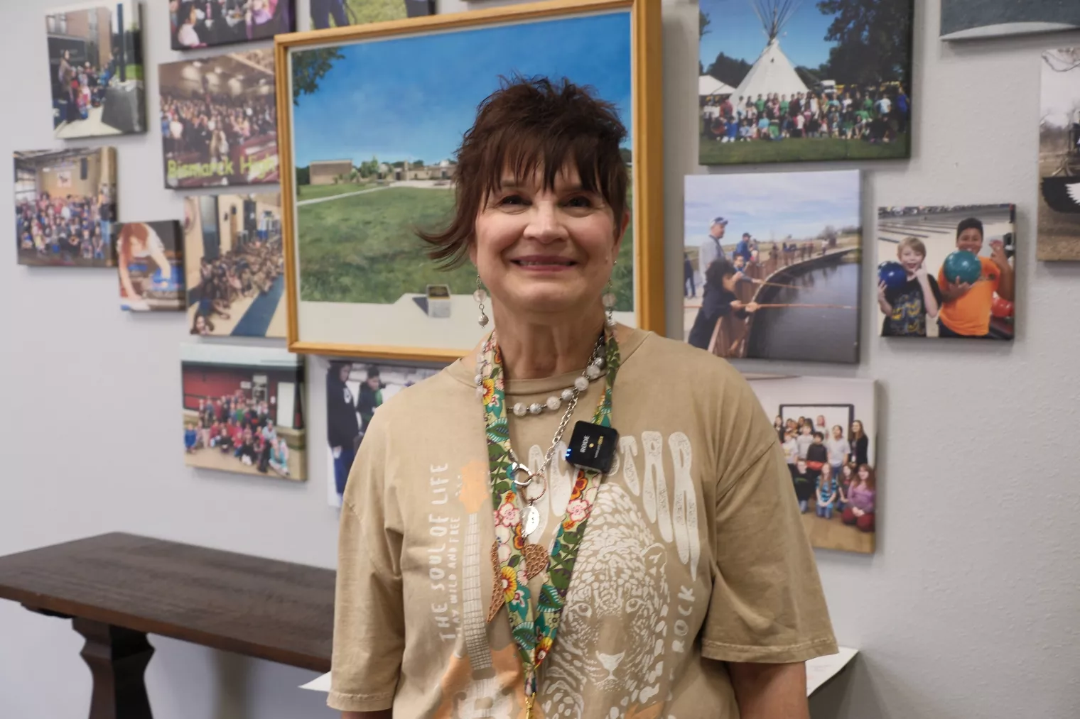 A woman with short brown hair smiles while standing in front of a collage of photos on a wall. She is wearing a beige shirt with a graphic and a necklace with multiple colorful pins. A framed picture of a park scene is prominently displayed behind her.