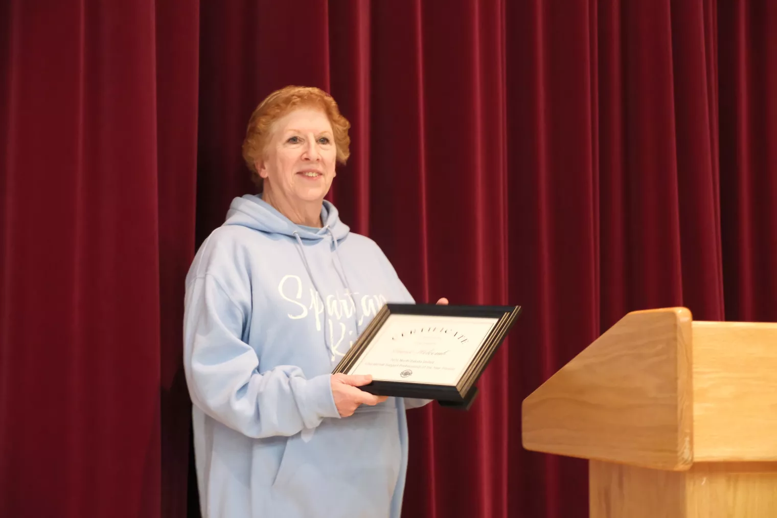A person in a light blue hoodie holding a framed certificate stands next to a wooden podium against a red curtain backdrop.