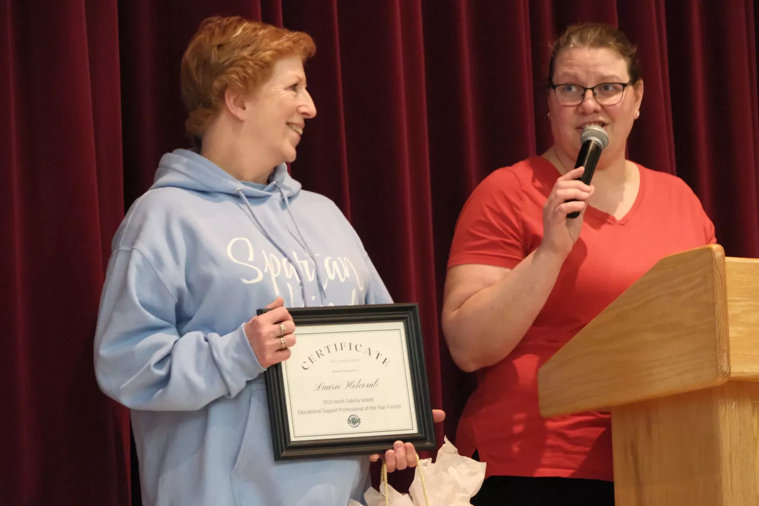 Two people stand on stage in front of a red curtain. One is holding a framed certificate and smiling, while the other speaks into a microphone from behind a podium.