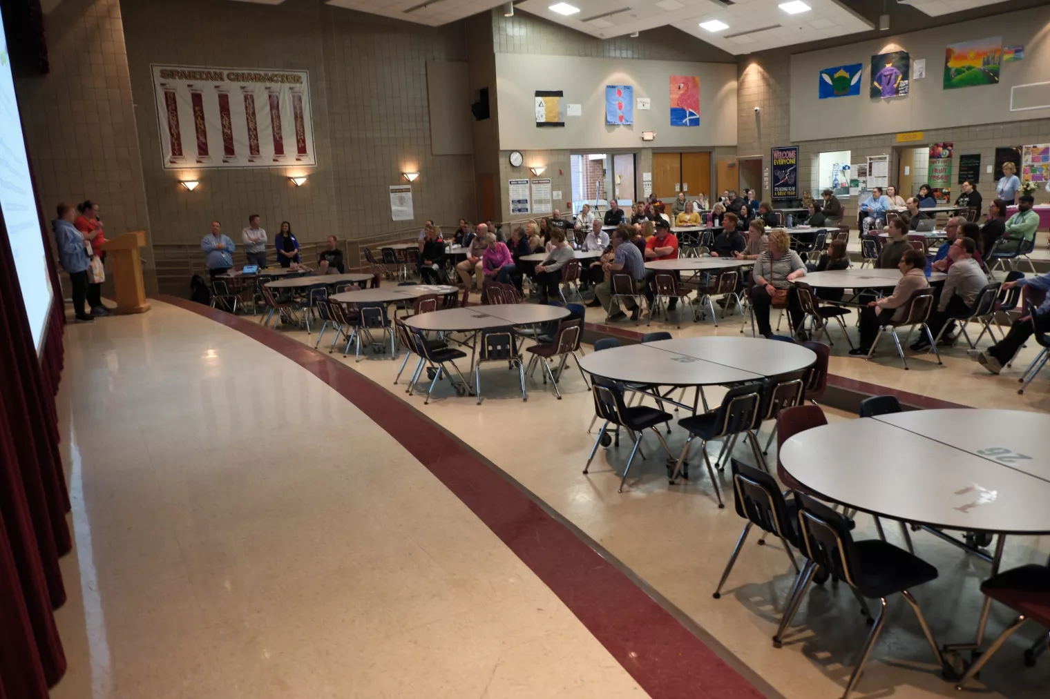A cafeteria with round tables and people seated, attending a meeting or presentation. A speaker stands at a podium to the left. Banners and school-themed decor are visible on the walls.