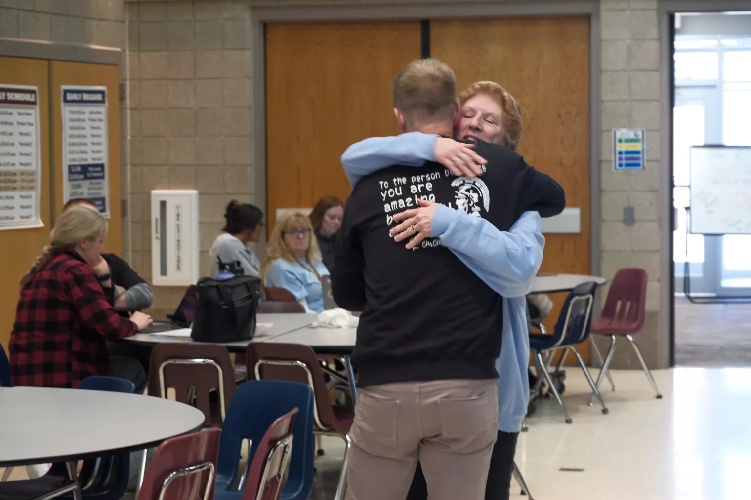 A person with a shirt reading "To the person ... you are amazing" hugs a woman in a communal room with other people seated at tables. The atmosphere is warm and casual, with soft lighting and neutral-colored walls.