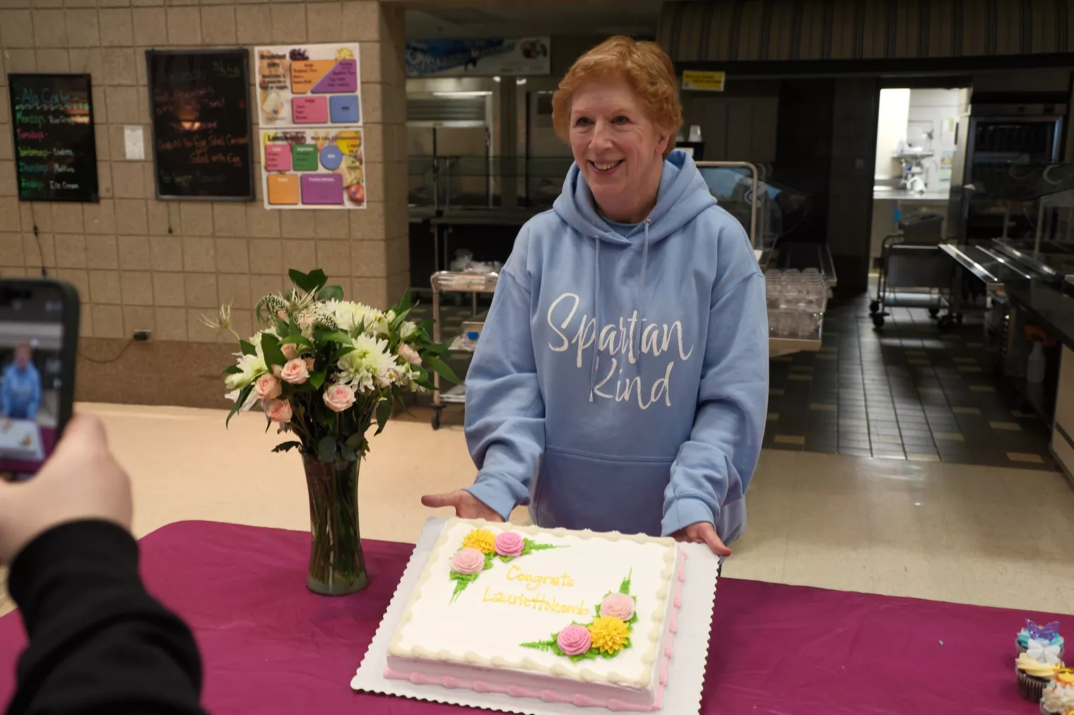 A person in a blue hoodie with "Spartan Kind" text stands behind a decorated cake and a bouquet of flowers. The cake reads "Congrats Laurie Holcomb!" as someone takes a photo. The setting appears to be a cafeteria.