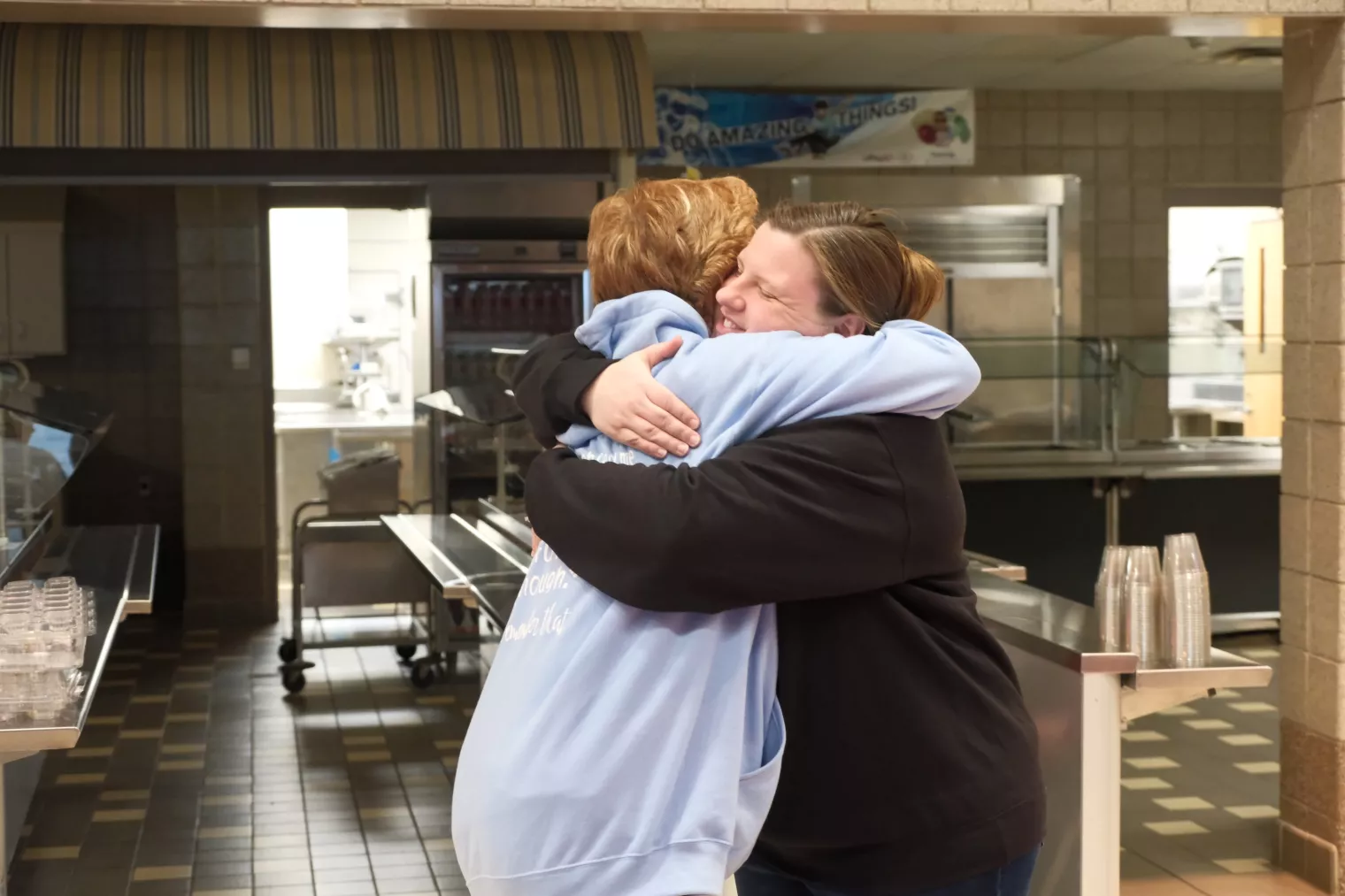 Two people are embracing in a kitchen area. One person is wearing a light blue hoodie, and the other is in a dark top. Their surroundings include stainless steel equipment and stacked plastic cups.