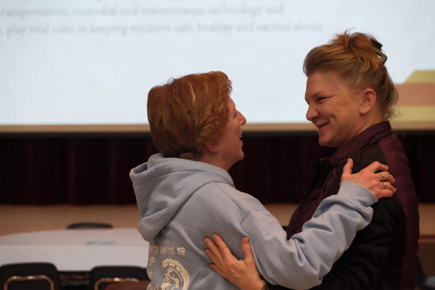 Two people stand facing each other, smiling warmly. One is wearing a light blue hoodie and the other a dark jacket. They appear to be in a spacious room with a projection screen in the background.