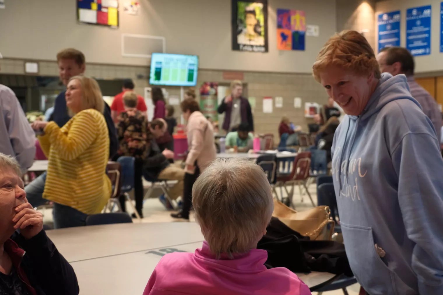 A group of people gathered in a communal space with tables and chairs. Two women are conversing, one standing and smiling, wearing a blue hoodie. Various people are in the background, some sitting and others standing near colorful signs.