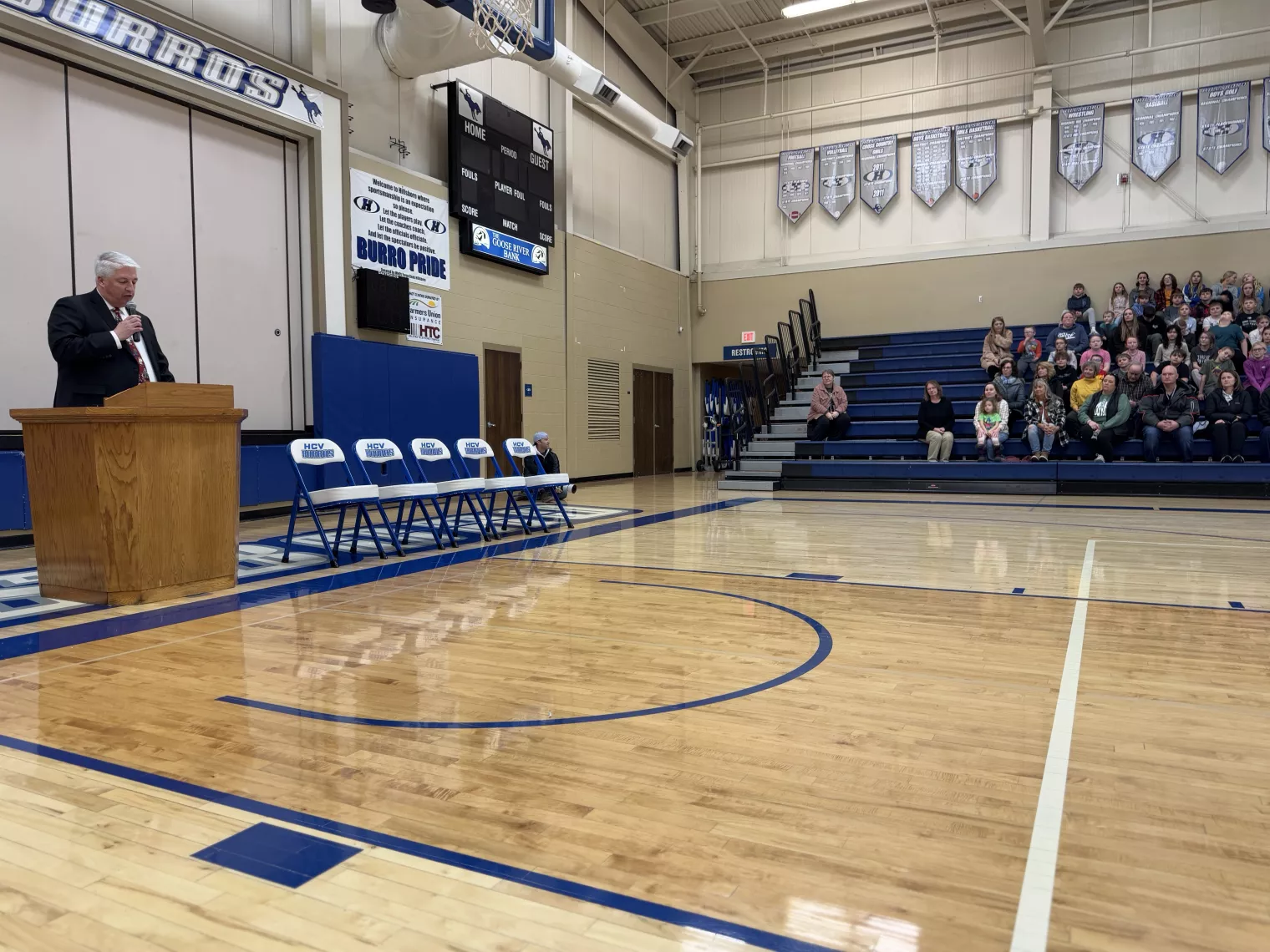 Hillsboro superintendent Jon Dryburgh speaking in front of a podium inside a school gymnasium that is filled with students and staff.