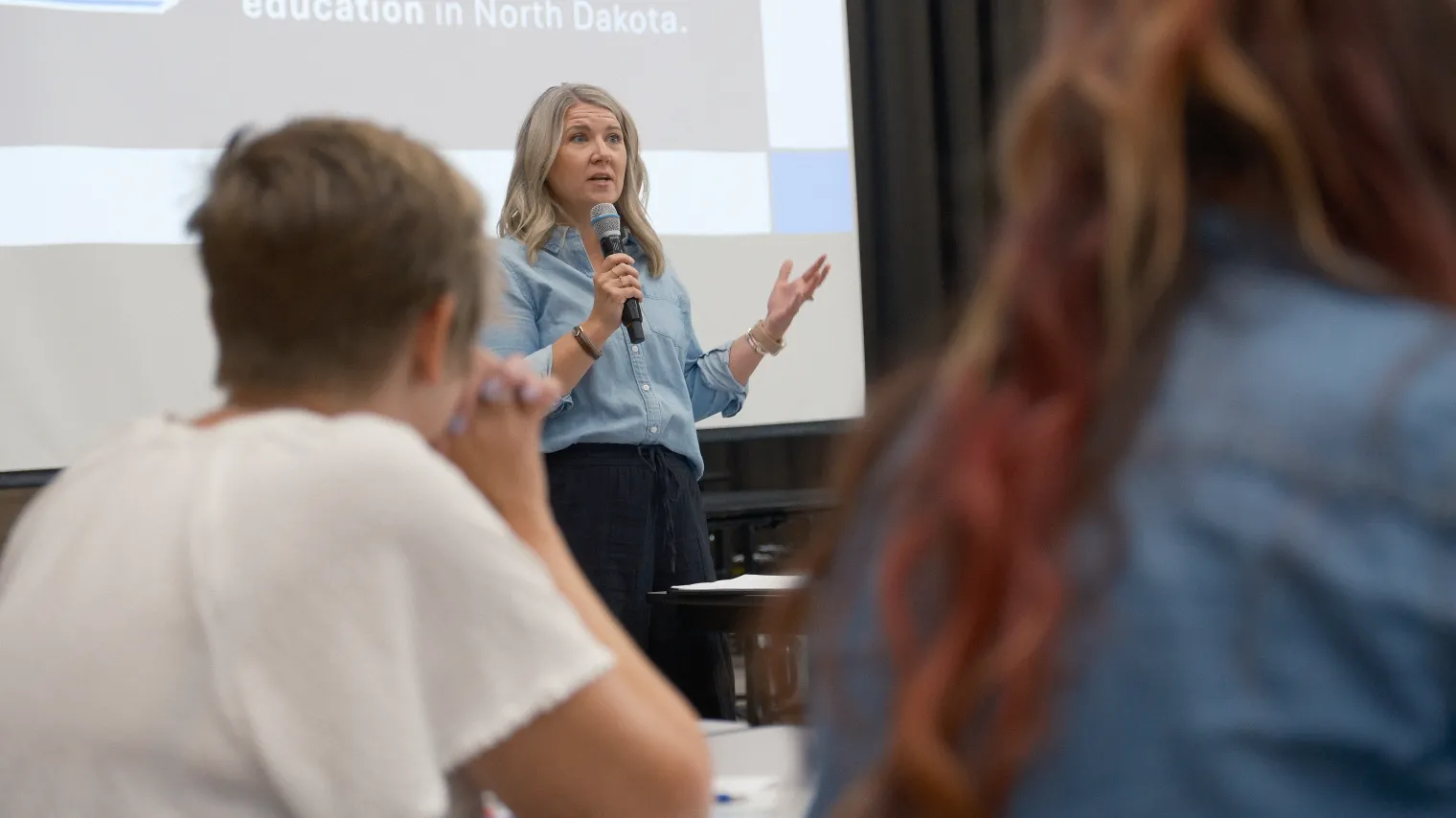 A person stands at front of crowd in school gymnasium, speaking into microphone.