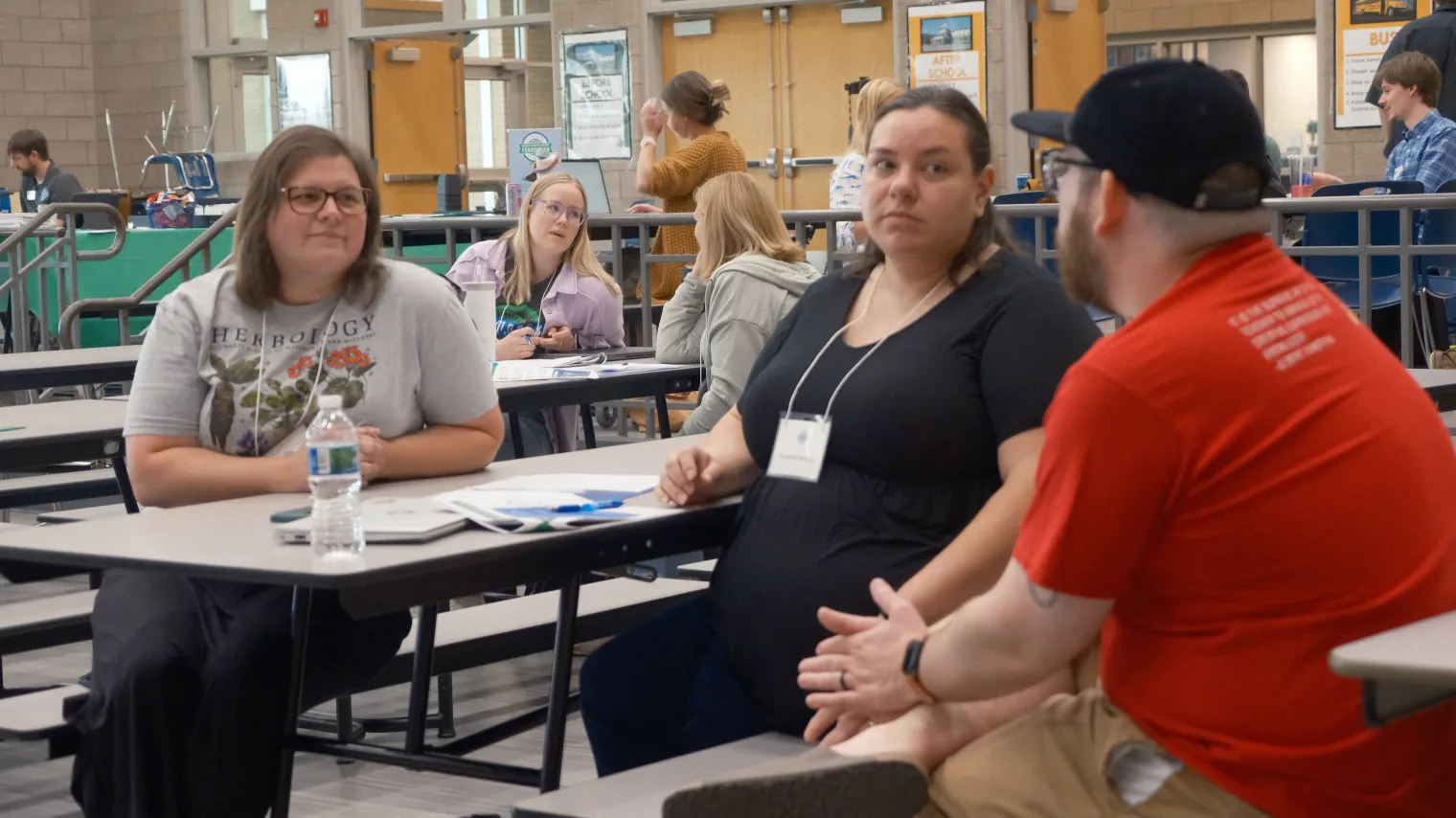 Two women and one man sitting at tables in school cafeteria, talking.