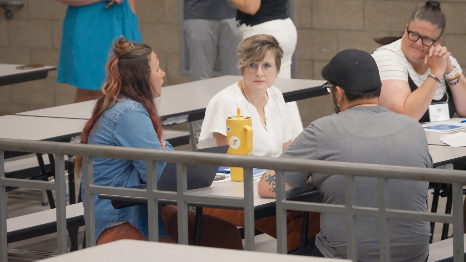 Group of people sitting around long table in school cafeteria, talking to each other.