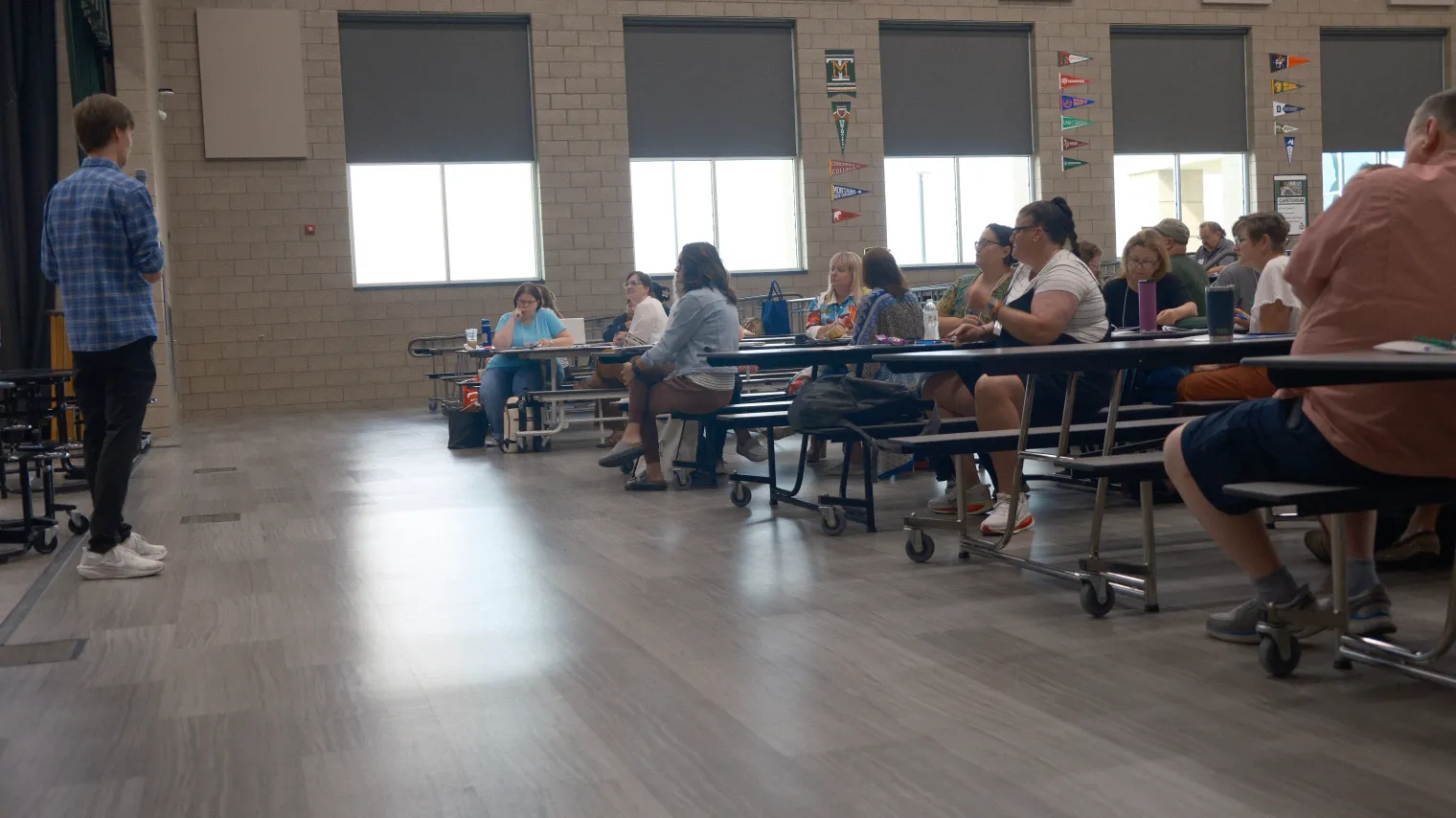 School cafeteria filled with people sitting at multiple tables, listening to a man speaking at front.