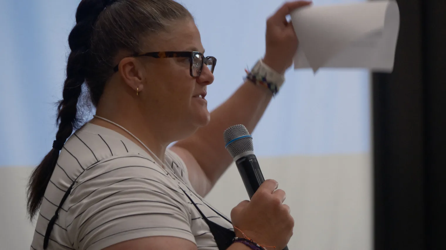 Woman with glasses and long hair braid, holds a microphone and piece of paper while talking to group.