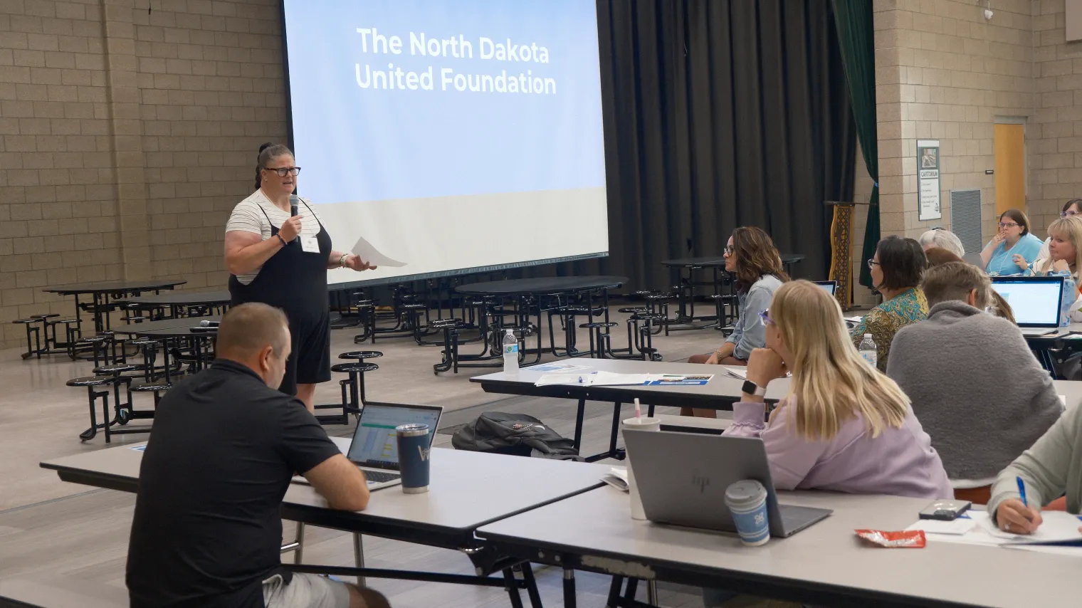 Woman standing in front of projector image that reads "North Dakota United Foundation" speaking to group of people in school cafeteria.