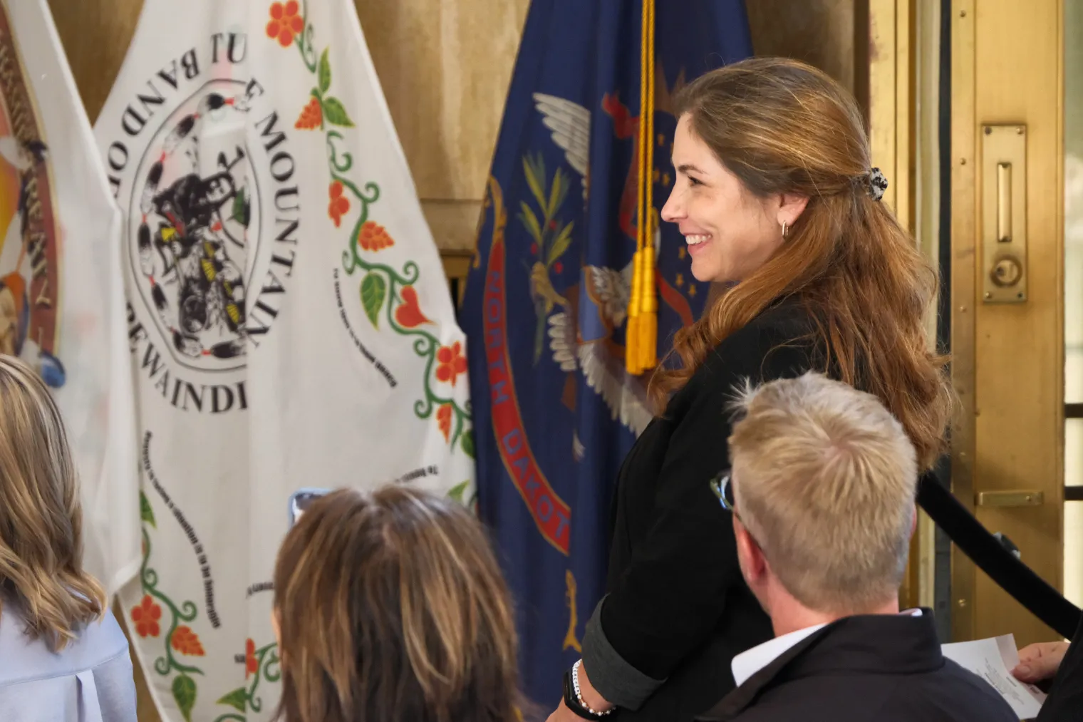 Woman with brown hair standing in hall of state Capitol among group of seated people.