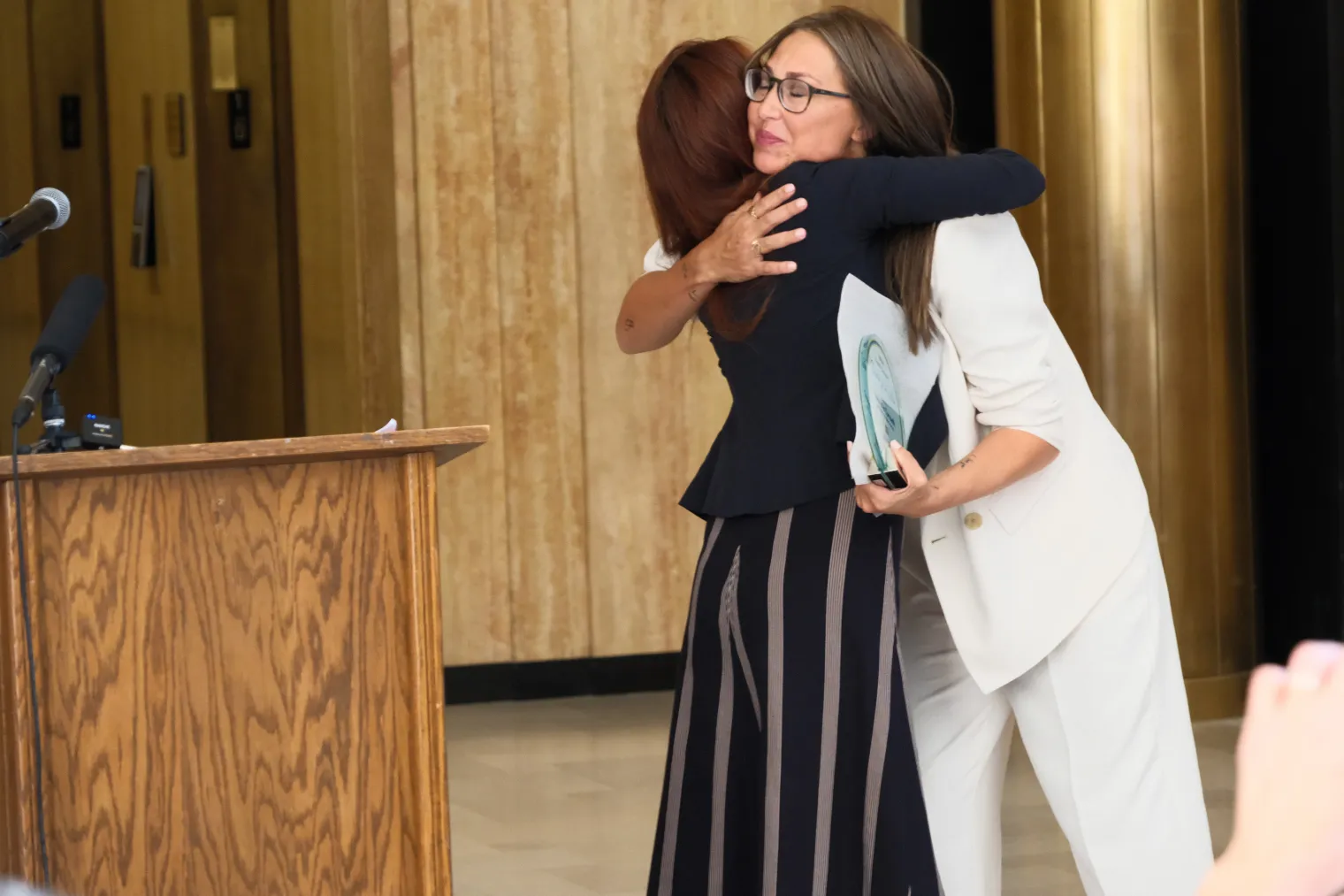 "A woman with long brown hair hugs another woman with glasses and straight brown hair, who is dressed in a white blazer and black top. 