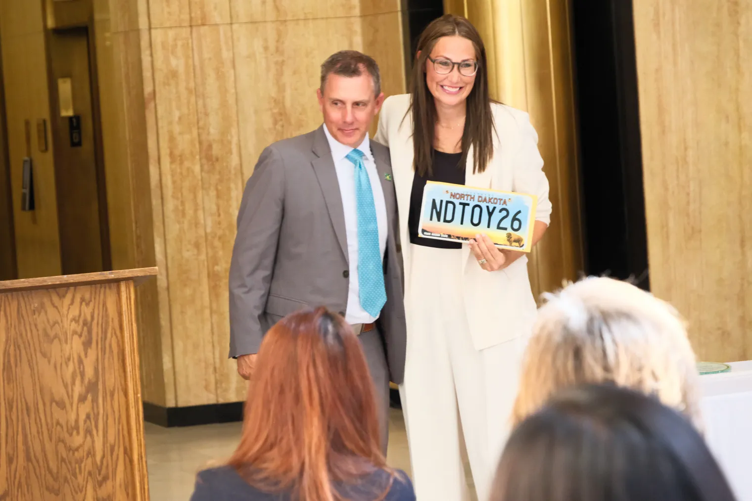 Woman in white suit standing next to man in gray suit, while she holds North Dakota license plate reading "NDTOY26."