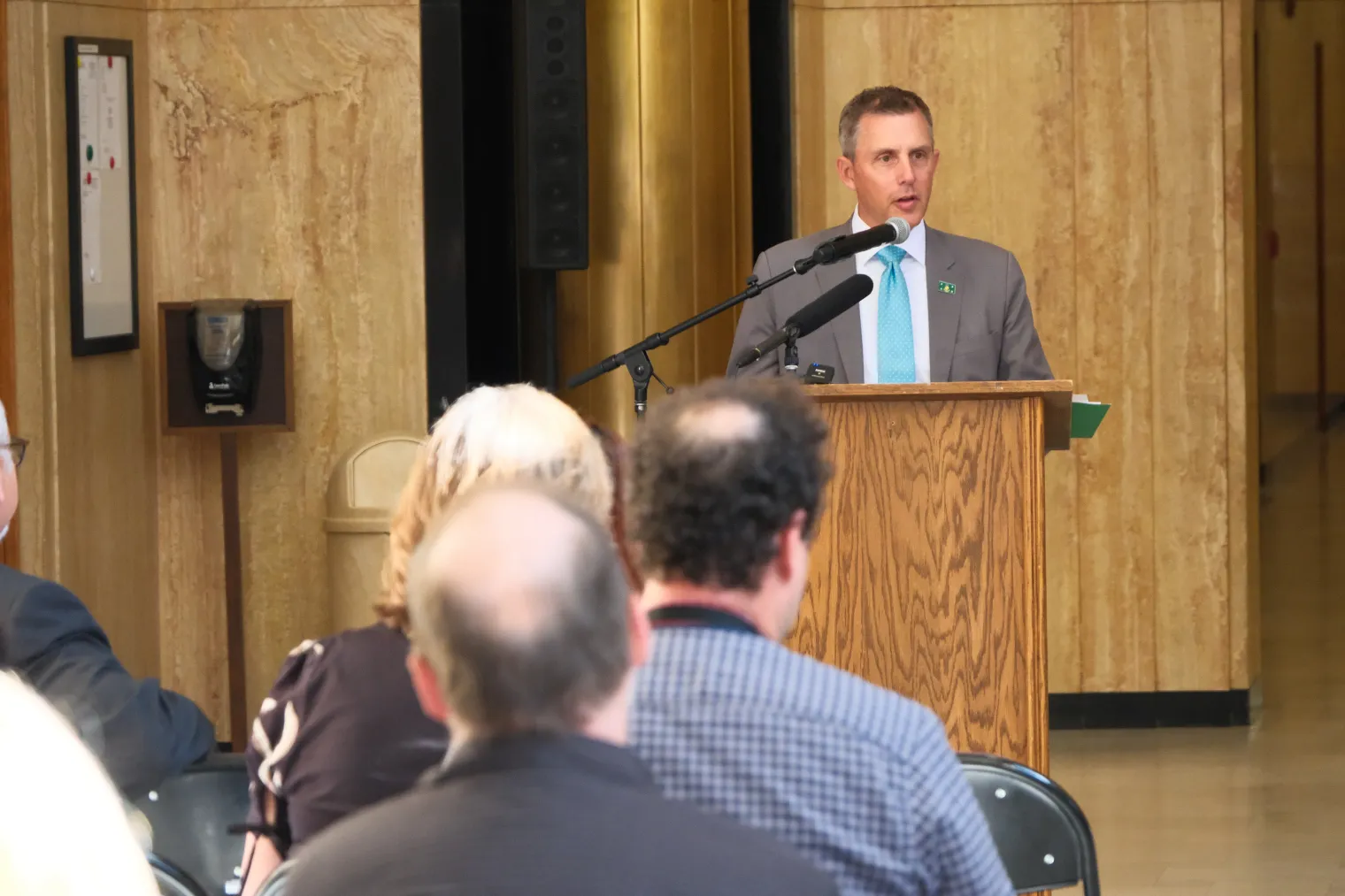 Man with short brown hair in gray suit with light blue tie and white shirt stands behind a podium, speaking to a group of people in a large hall.