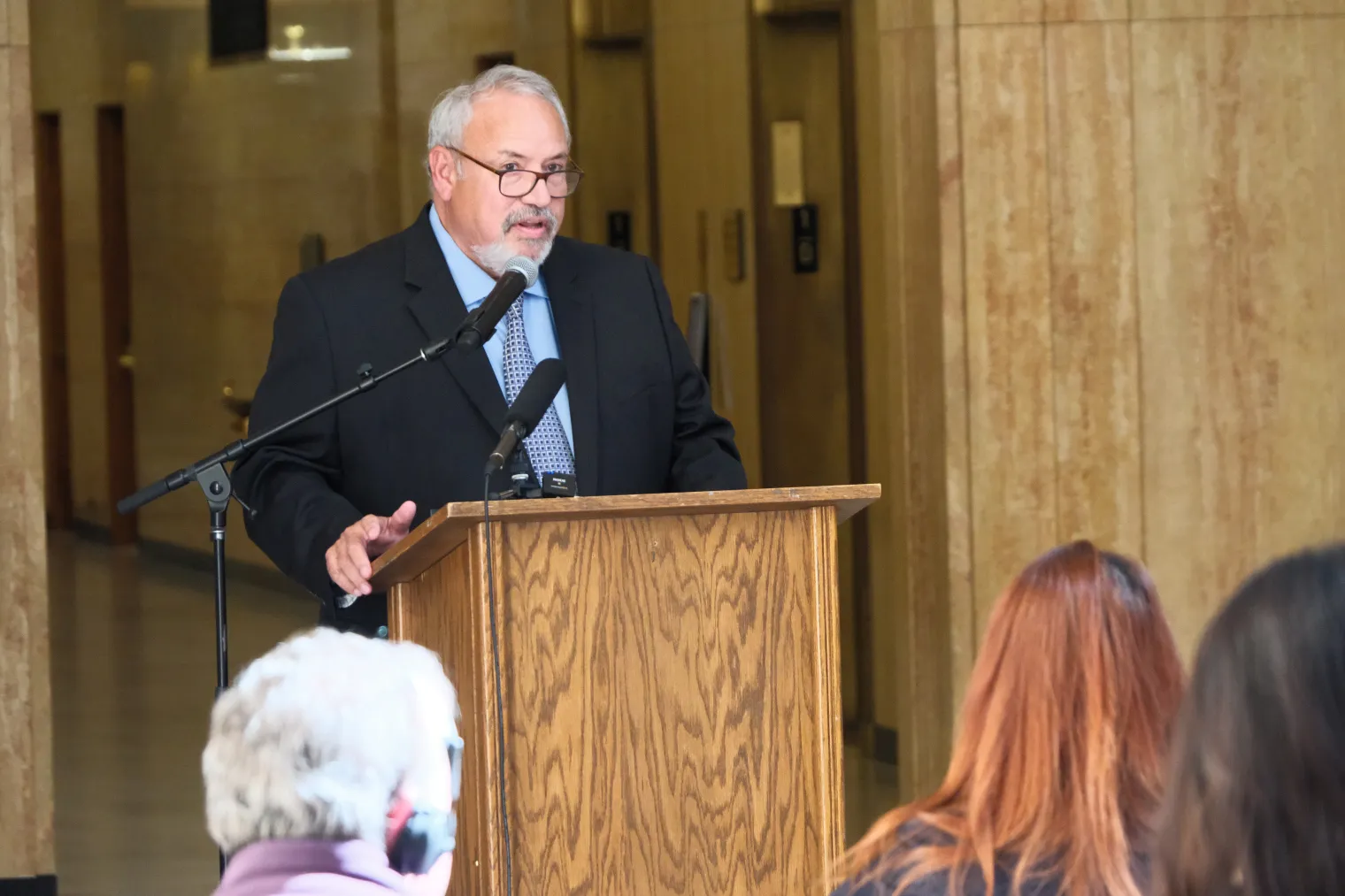 A man in glasses with short, gray hair and glasses speaks into a microphone from behind a podium in a large hall. 