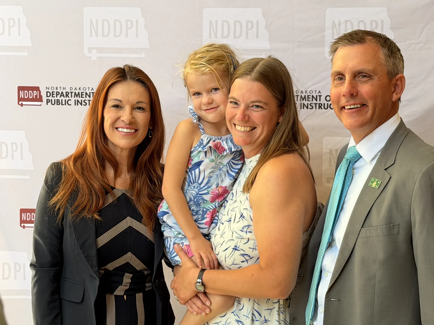 A woman with long brown hair in black dress standing next to woman with long blonde hair in white dress, holding a young girl in blue dress, with man in gray suit, white shirt and light blue tie, all are smiling.