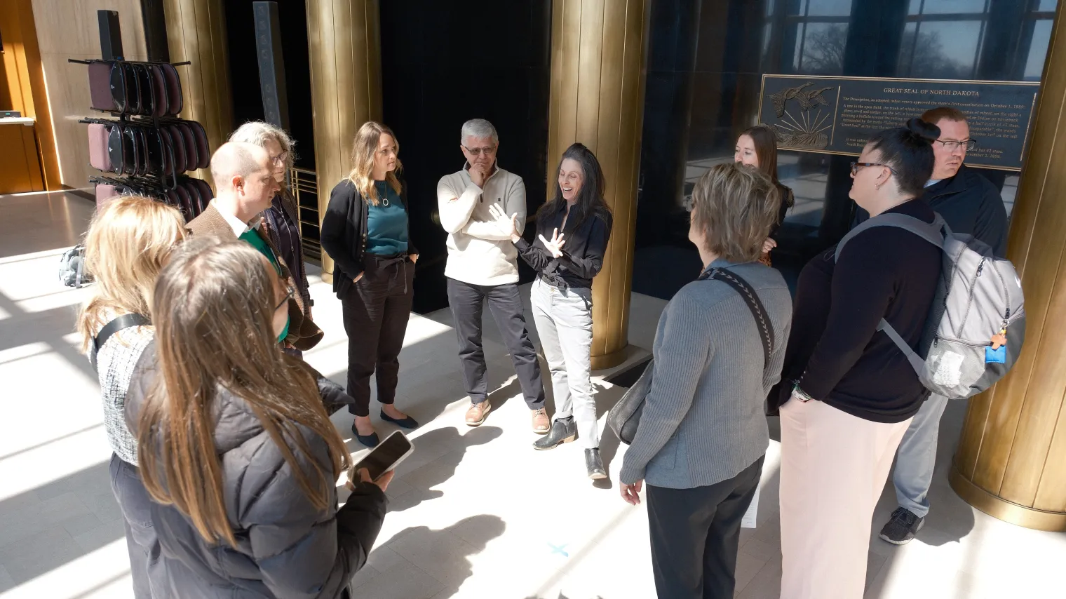 A group of North Dakota State Teachers of the Year (NDSTOY) chapter members stand in a circle inside the North Dakota Capitol, listening as one person speaks and gestures. Sunlight fills the space with tall columns and a state seal plaque visible in the background, suggesting a conversation or strategy discussion during an advocacy visit.