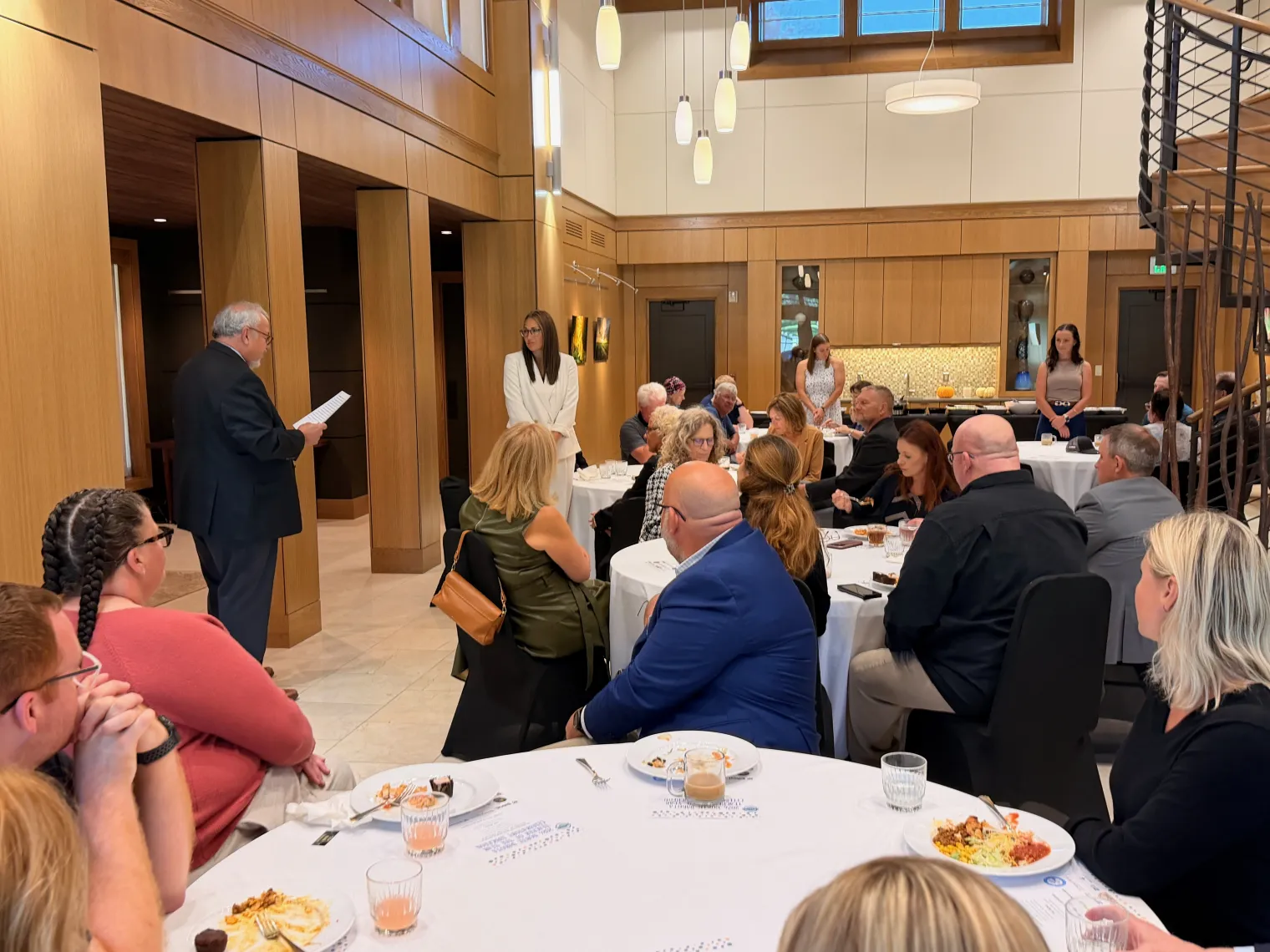 An audience of educators and guests sit at round tables during a luncheon in a wood-paneled event space. A man at the front reads from a paper while attendees listen, some with plates and glasses in front of them, suggesting a formal program or recognition ceremony.
