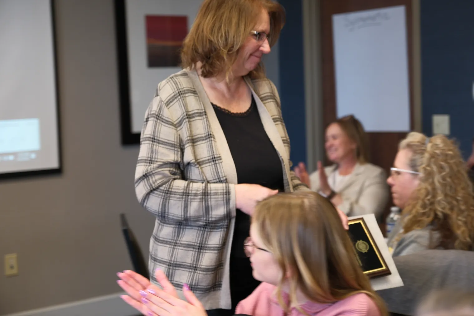 A woman wearing a black shirt and tan Cardigan sweater is applauded by group as she receives award.