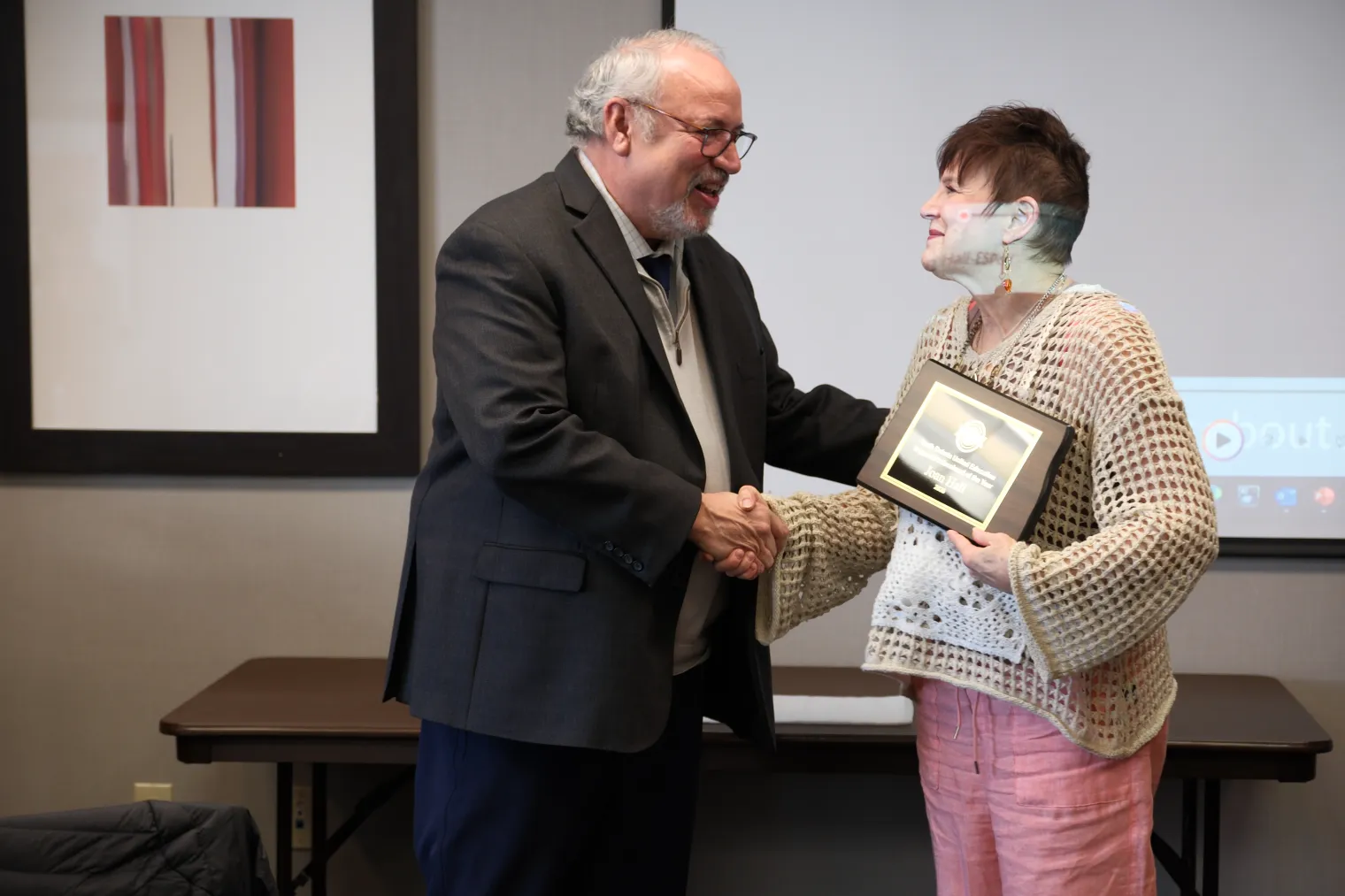 A man in suit and tie shakes hands with woman with shorter hair, who is holding an award plaque in the shape of the state of North Dakota.