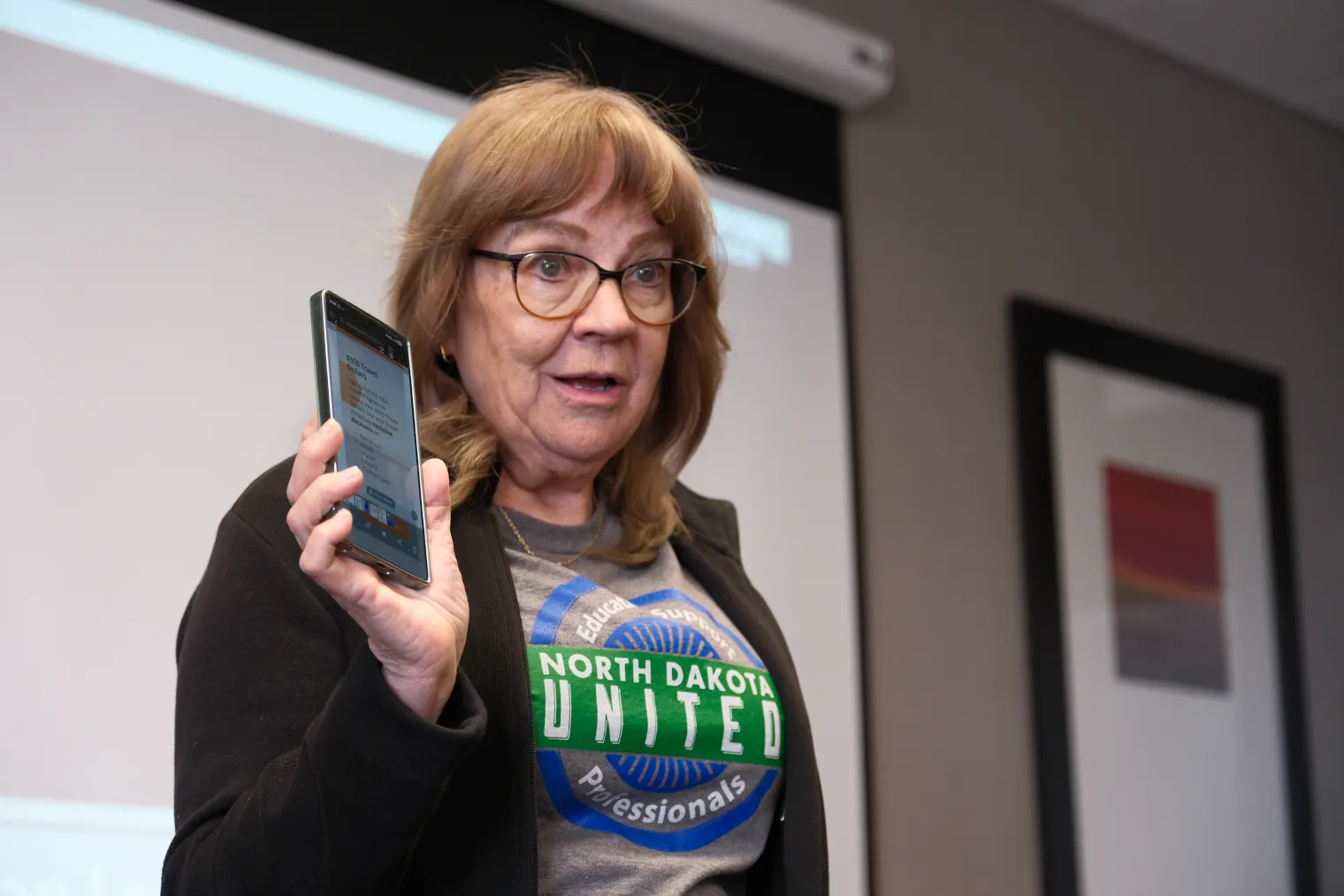 A woman wearing glasses, black jacket and gray shirt with the North Dakota United logo, holds a cell phone while speaking to group.
