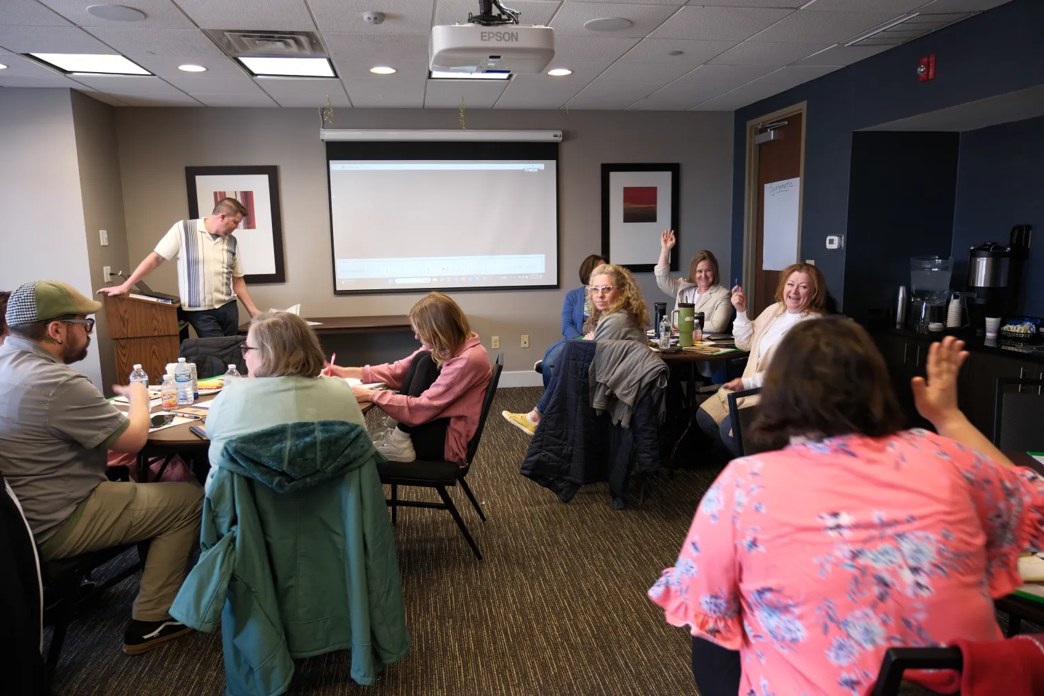 Conference room filled with people sitting around tables, listening to speaker at front of the room.